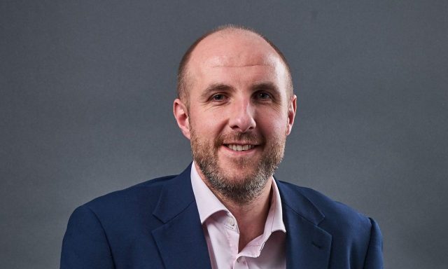 James Bradshaw standing in front of grey screen wearing blue suit and pink shirt