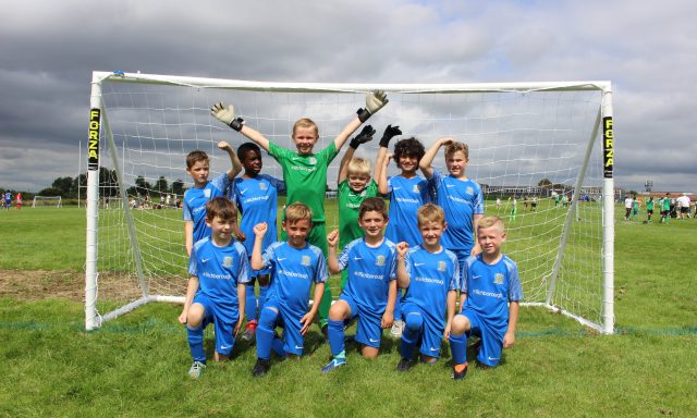 Picture shows an under nine year-olds boys football team, posing in a goal in their new blue kits with the goalie in his green kit in the middle. They are all cheering and smiling at the camera.