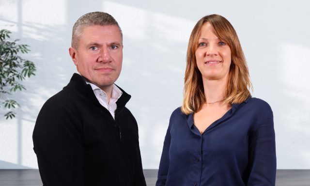 Image shows a smiling white man and woman in smart casual dress standing side by side.
